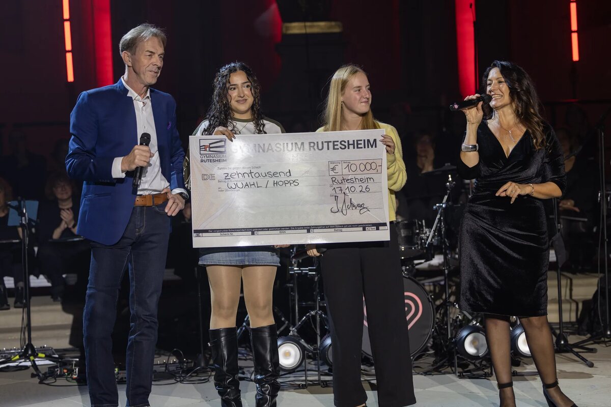 Schulleiter Jürgen Schwarz und zwei Schülerinnen des Gymnasiums Rutesheim mit dem großen Spendenscheck über 10.000 Euro auf der Bühne der Friedenskirche, daneben die Moderatorin